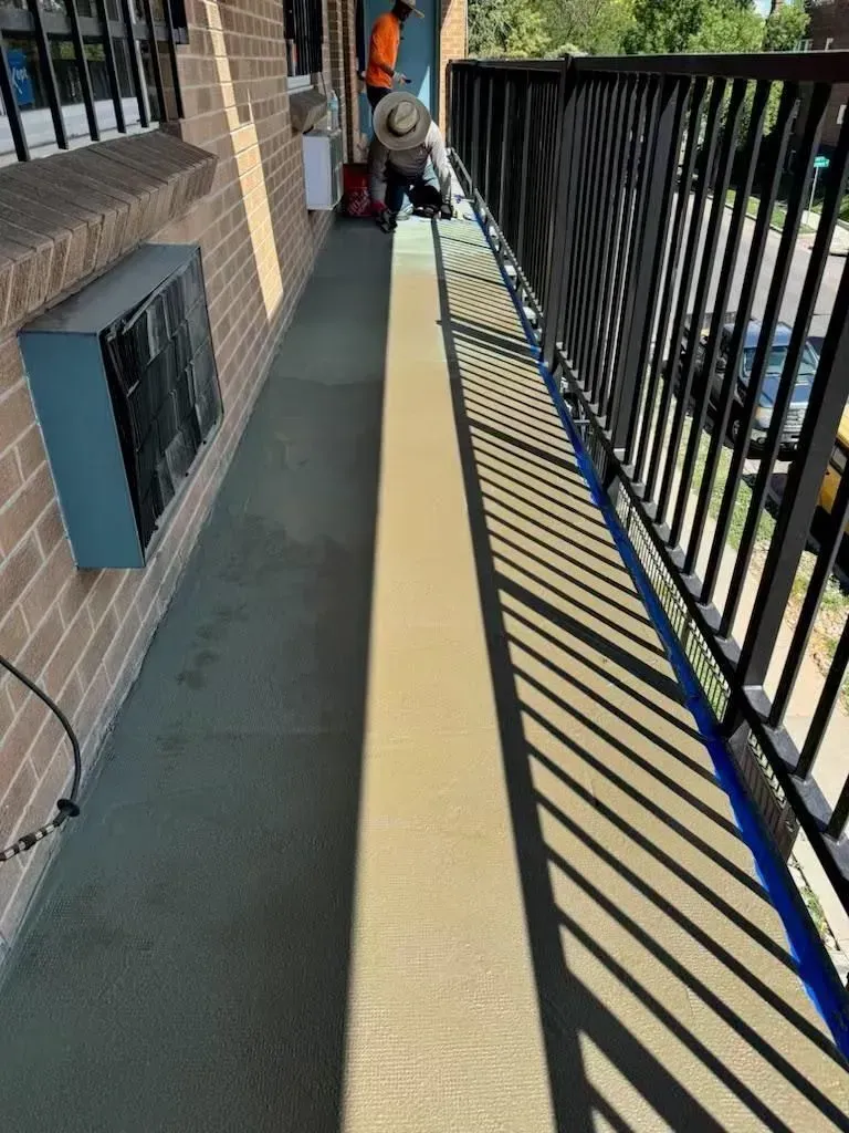 A person is grinding a balcony floor. The balcony has a black railing and is next to a brick building.