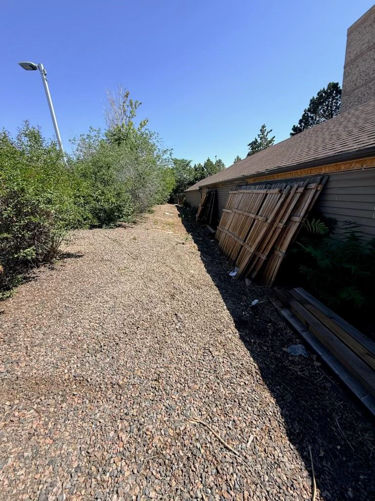 Gravel pathway beside a building with stacked boards, shrubs, and a clear blue sky