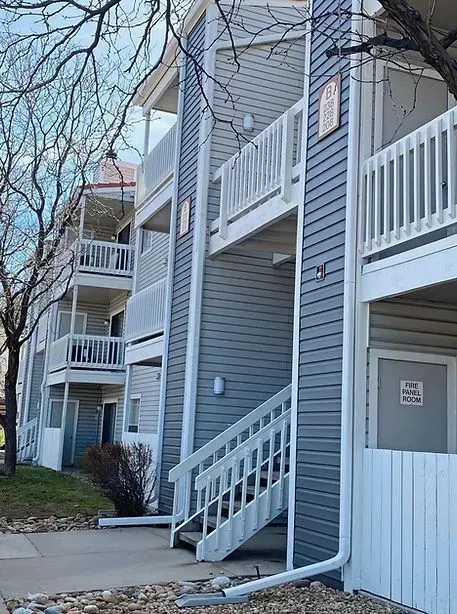 Apartment building with gray siding, white stairs, and balconies; a bare tree is in the foreground.