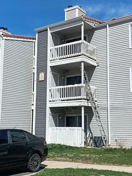 Apartment building with a ladder propped against a balcony. Gray siding, sunny day, and a car parked below.