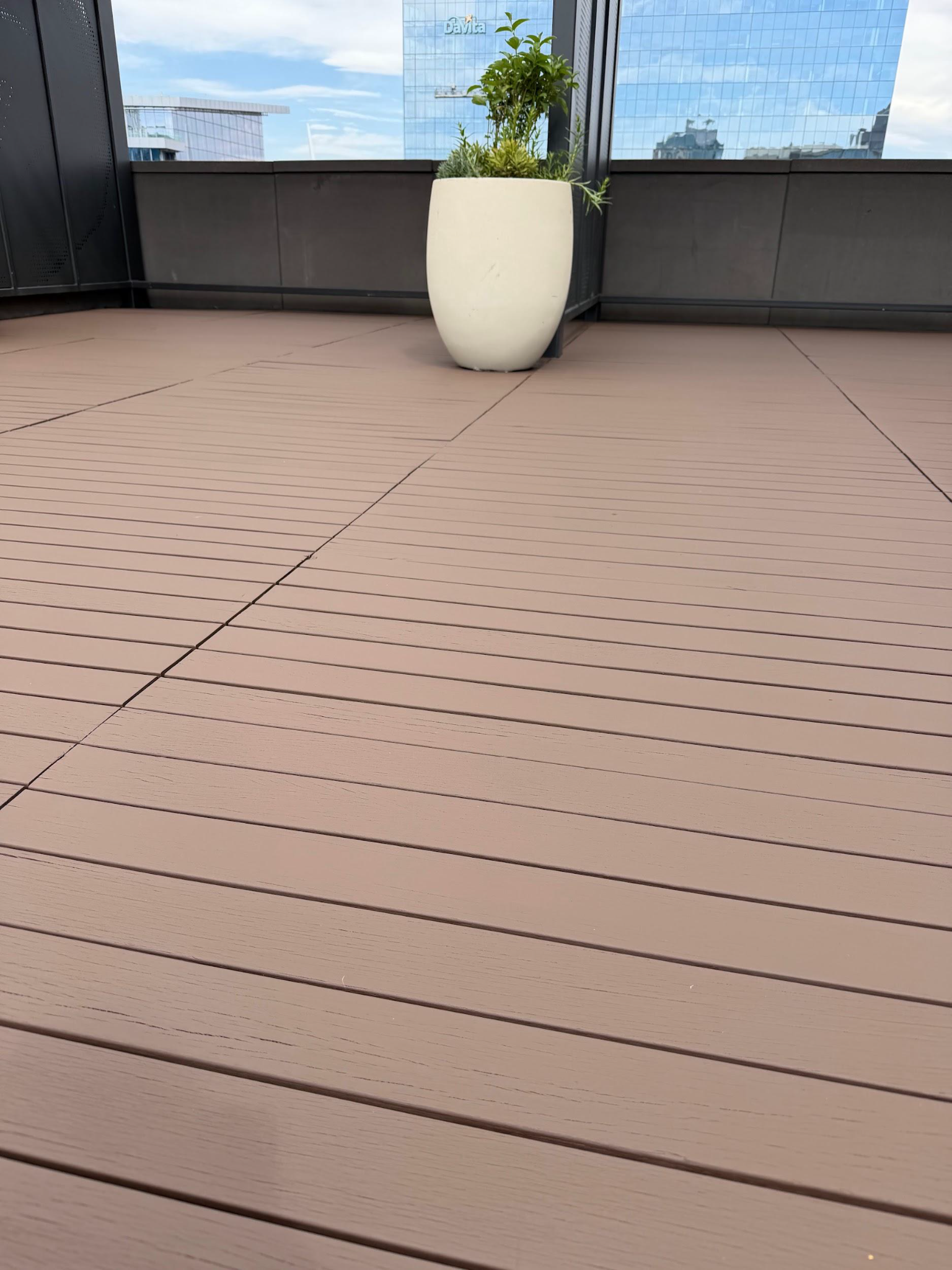 Empty rooftop deck with brown flooring and a white planter near glass railing, city skyline in background