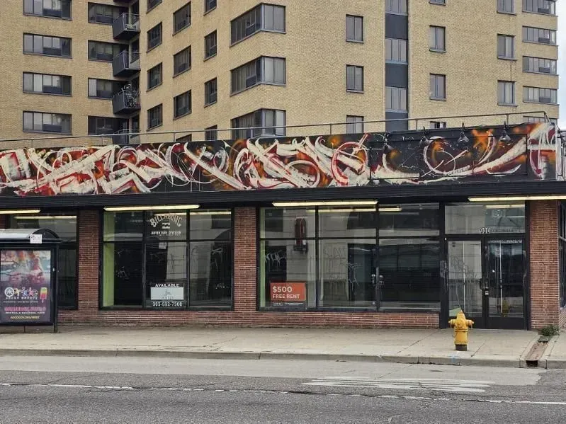 Storefront with abstract mural, brick facade, and a bus stop in front of a tall building.