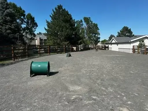 Green dog agility tunnel in a gravel dog park, with a fence and houses in background.