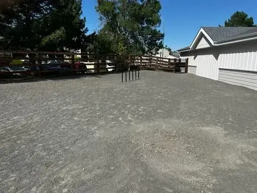 Gravel lot with fence, gray building, trees, and poles. Sunny day.