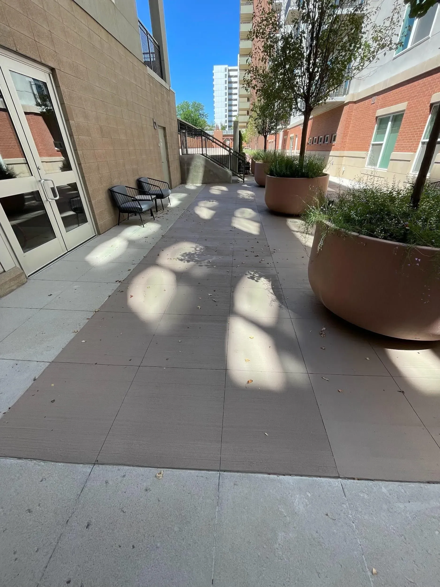 Narrow outdoor walkway with planters and a building on each side. Bright sunlight creates shadows.
