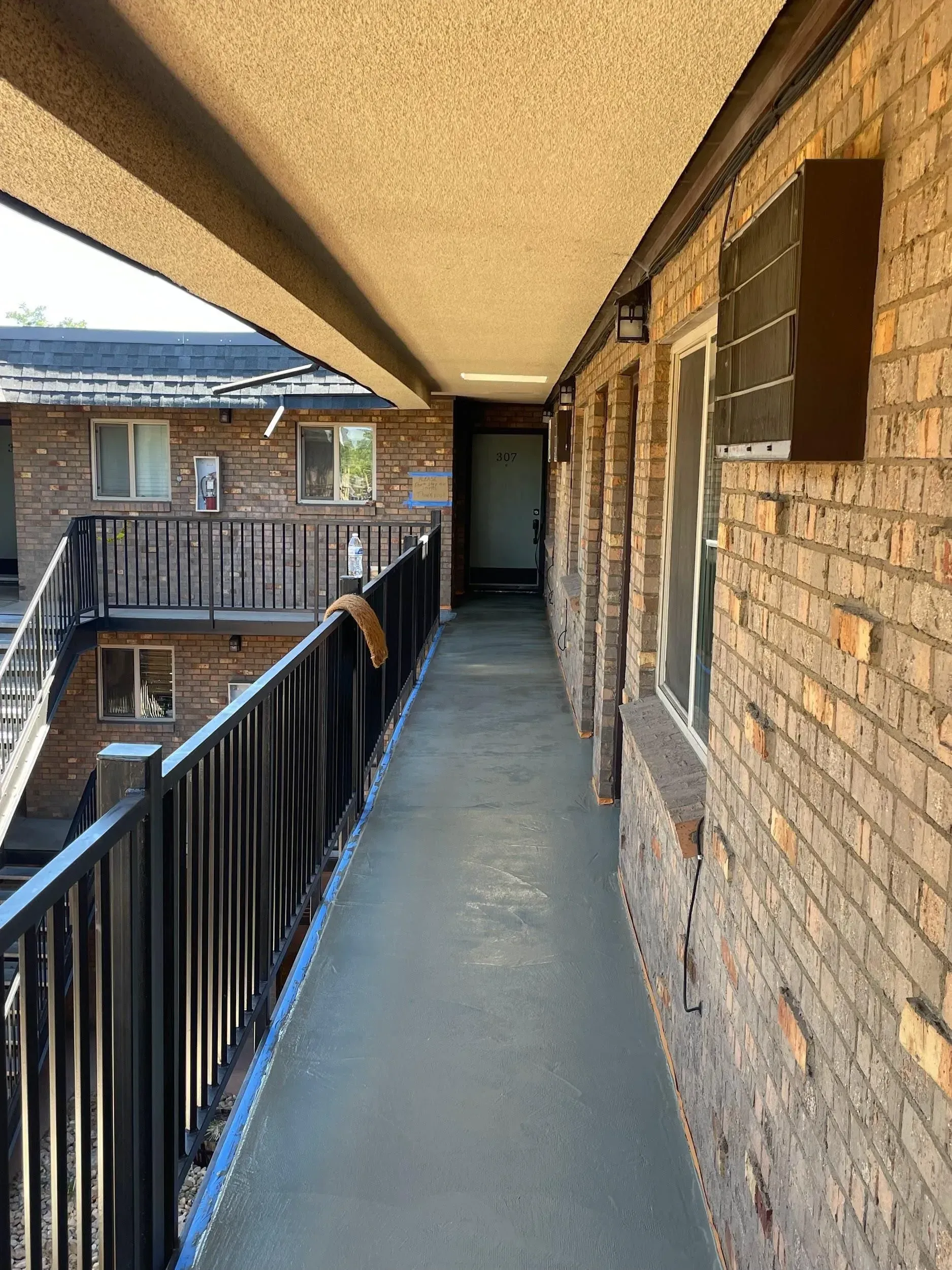 Exterior apartment hallway with brick walls, doors, and black railing.