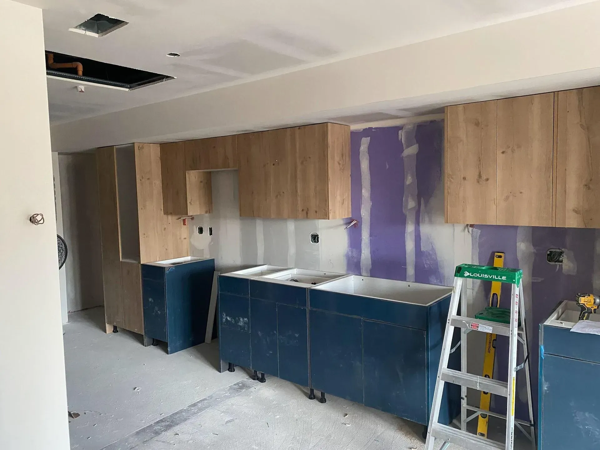 Kitchen under construction: blue cabinets, wooden wall cabinets, drywall, ladder, and unfinished ceiling.