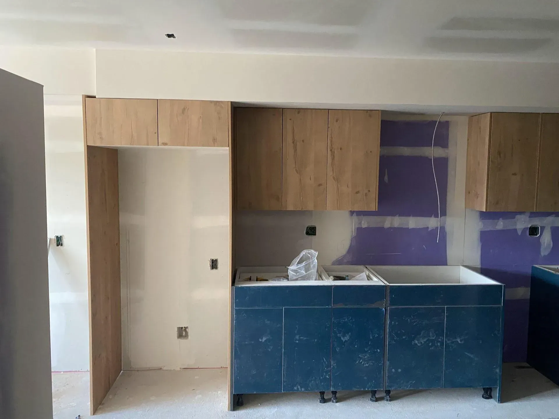 Kitchen under construction: blue cabinets, wooden fridge surround, upper cabinets, drywall.