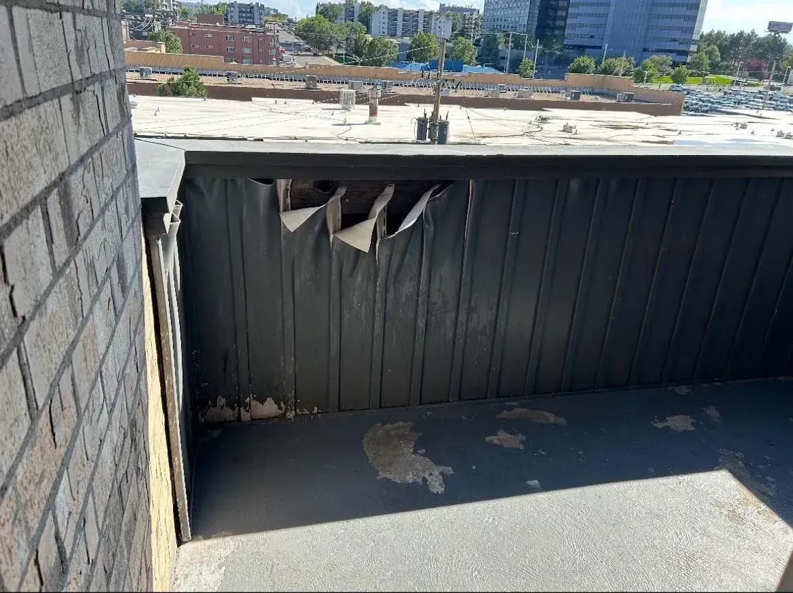 Exterior view of a rooftop with weathered black siding, revealing structural damage. Brick building in the background.