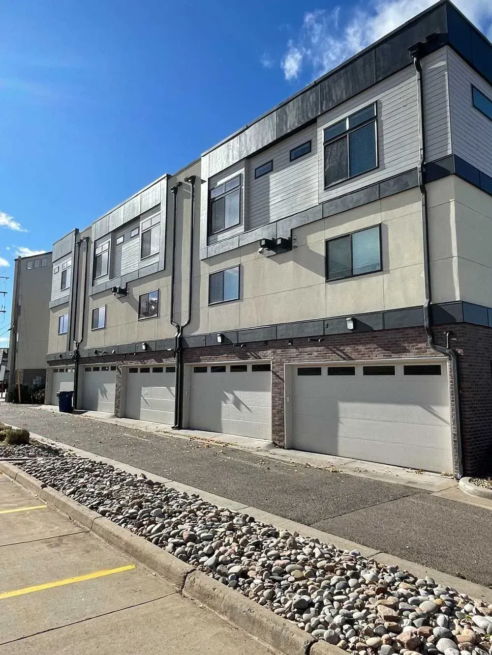 Row of townhouses with white garage doors and light-colored siding, set on a gravel drive under a blue sky.