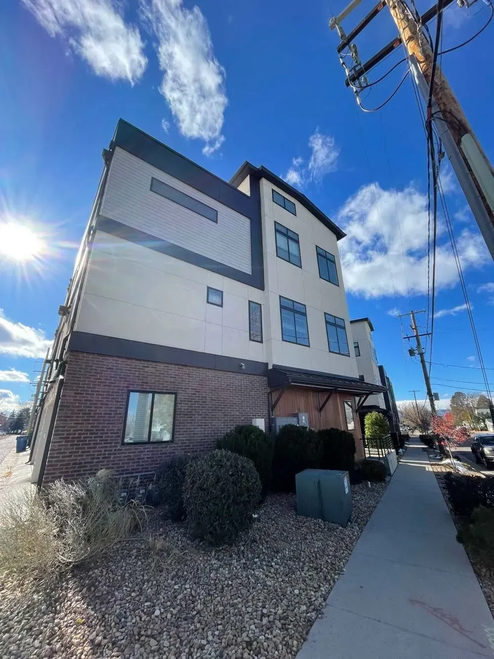 Multi-story modern building with brick base, beige and brown siding, blue sky, and sidewalk.