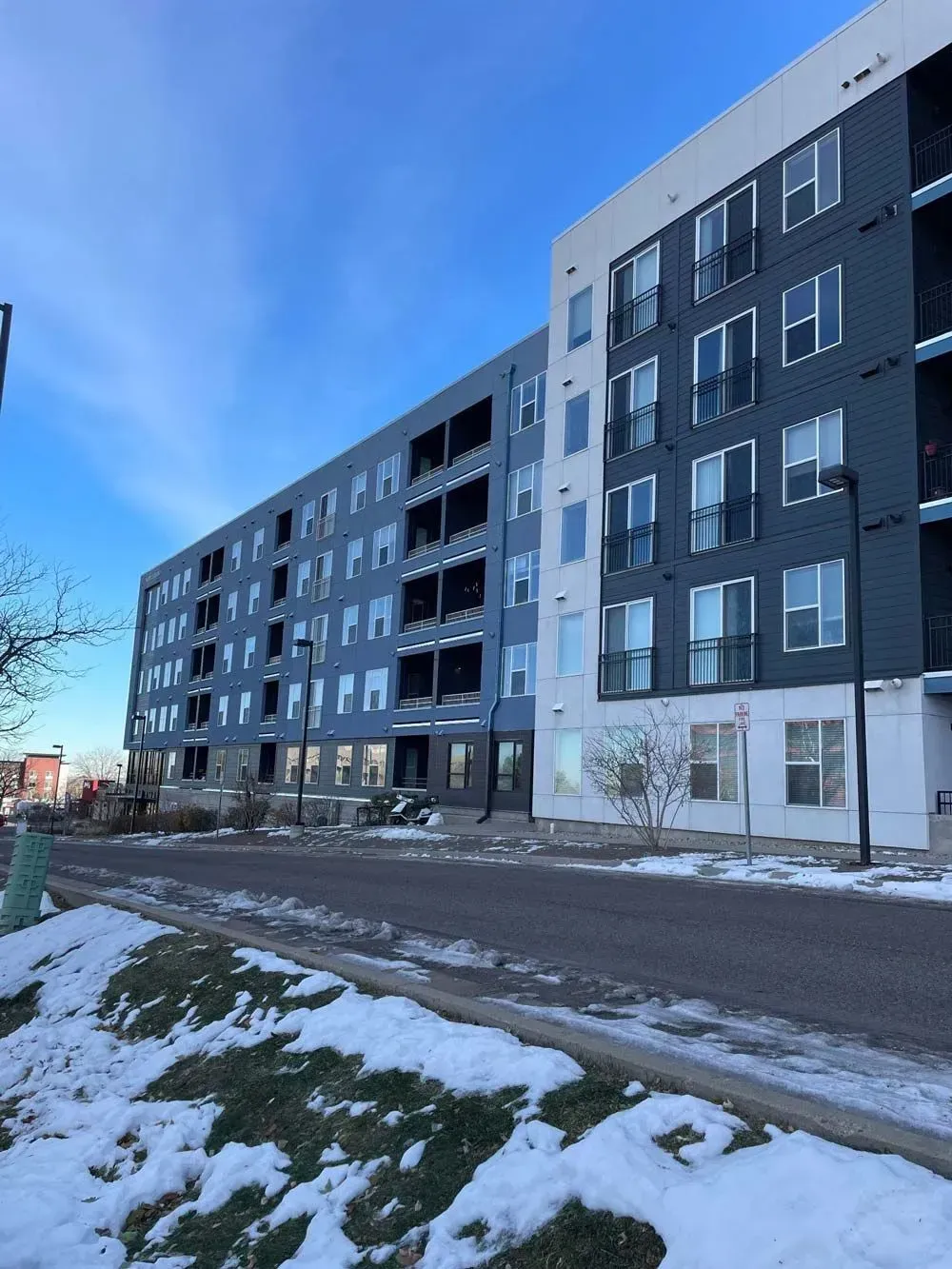 Modern apartment building with dark blue exterior and white accents, snow on the ground, blue sky.