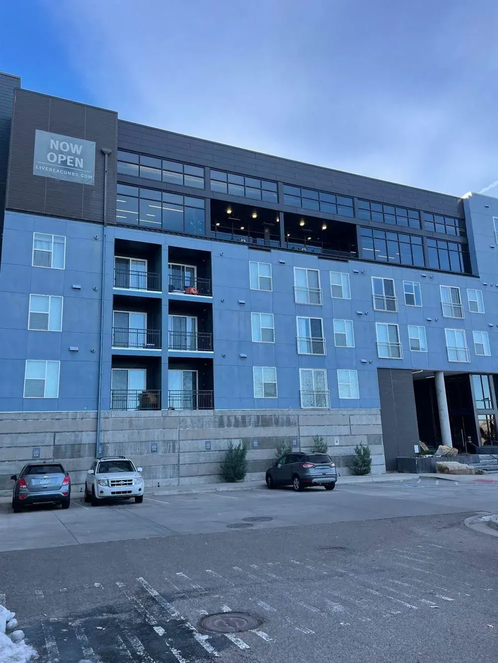 Modern blue apartment building with balconies, parked cars, and cloudy sky.