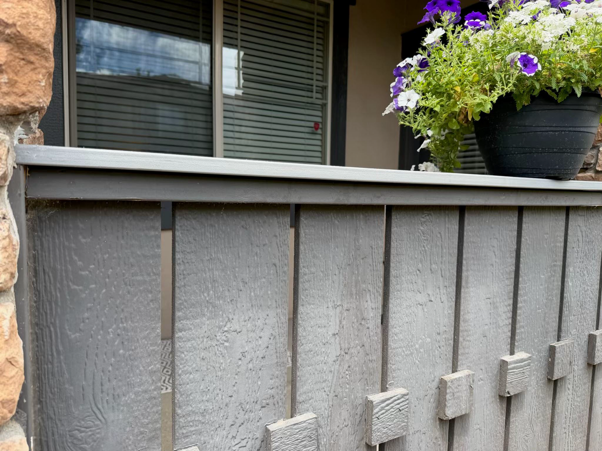 Gray porch railing beneath a window with blinds, topped by a black planter of purple and white flowers