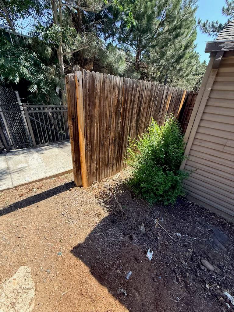 Backyard corner with a reed privacy fence, gravel ground, shrubs, and a house wall in sunlight