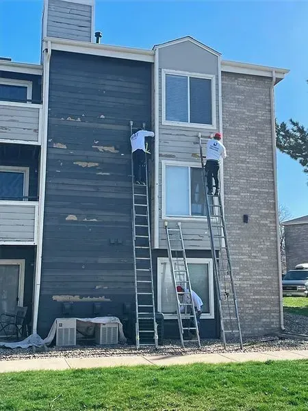 Three workers painting building exterior with long ladders, blue sky.