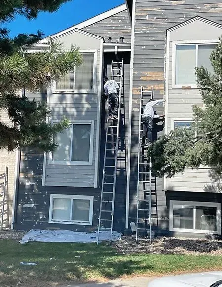 Two workers on tall ladders painting a gray building exterior.