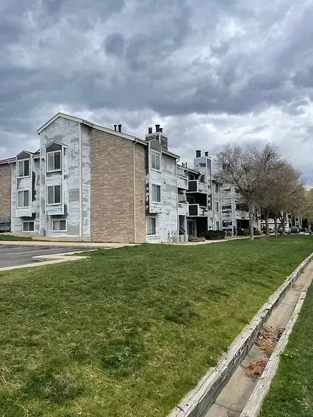 Apartment building with white siding and brick facade, next to a grassy area with a concrete ditch, under a cloudy sky.