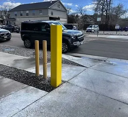 Sidewalk with bollards; a yellow square one and two brown cylindrical ones; car in the street.