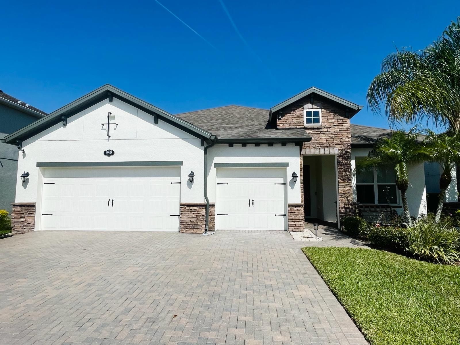 Two-car garage home with brick accents, blue sky. Paved driveway, green lawn, palm tree.