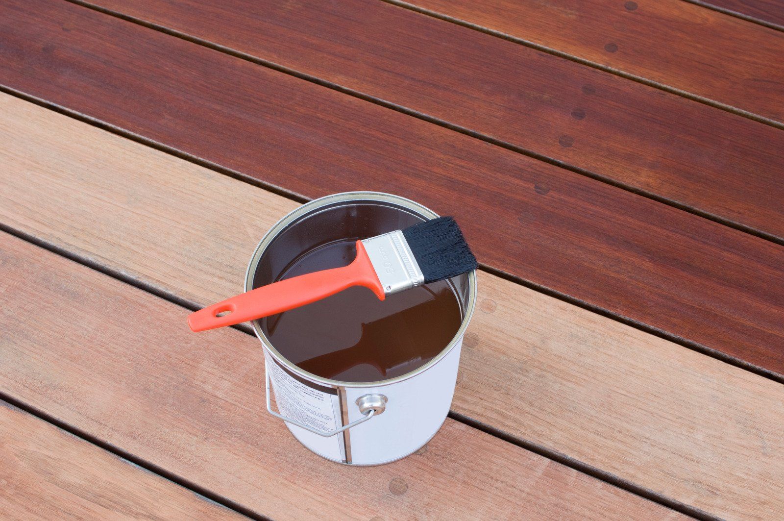 Paint can with brush on wooden deck, partly stained dark brown, partly unstained light brown.