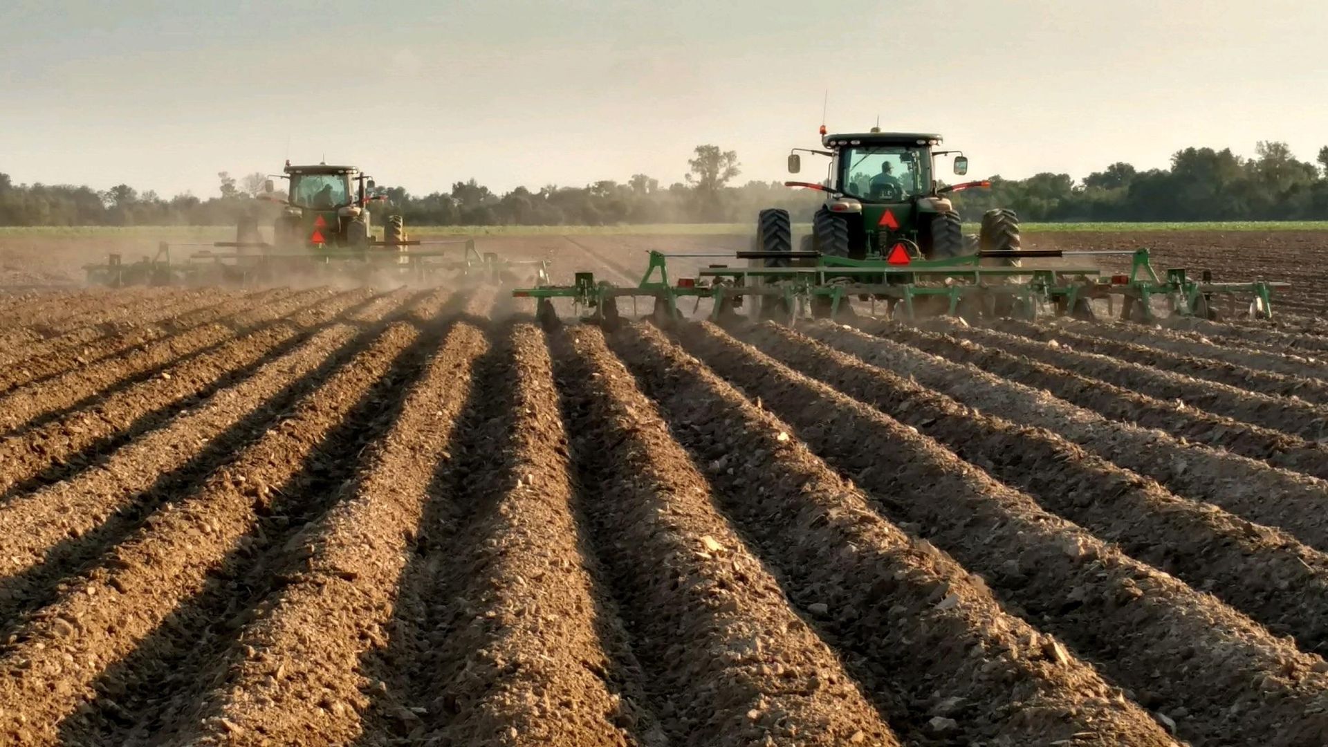 Two tractors are plowing a field with rows of dirt