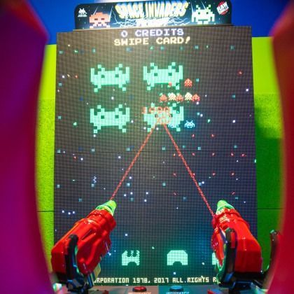 Hands firing red lasers at a classic Space Invaders arcade screen with green aliens and score text