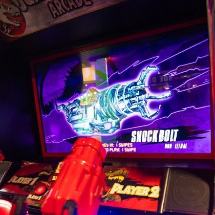 Arcade game screen showing a glowing blue rocket bolt target on a purple background, with a red gun in foreground