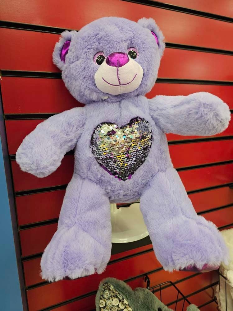 Lavender teddy bear with a sequined heart on display against a red slat wall