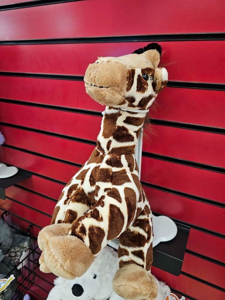 Plush giraffe toy on a shelf against a red slatwall, with a small white stuffed animal below.