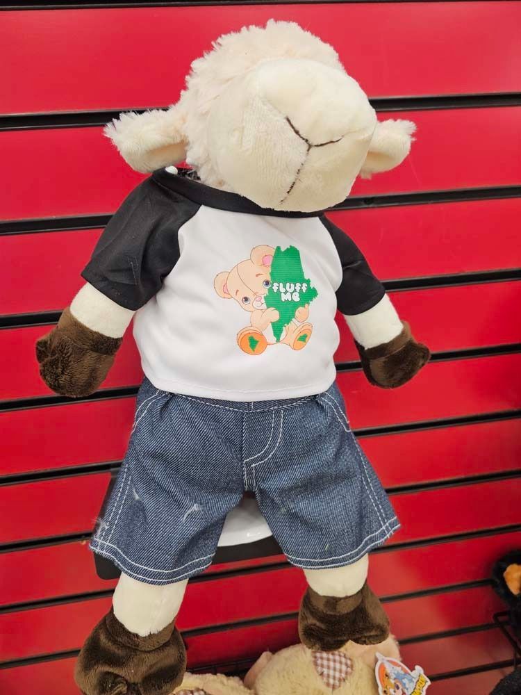 Plush sheep toy wearing a black-and-white shirt and denim shorts against a red slatted background