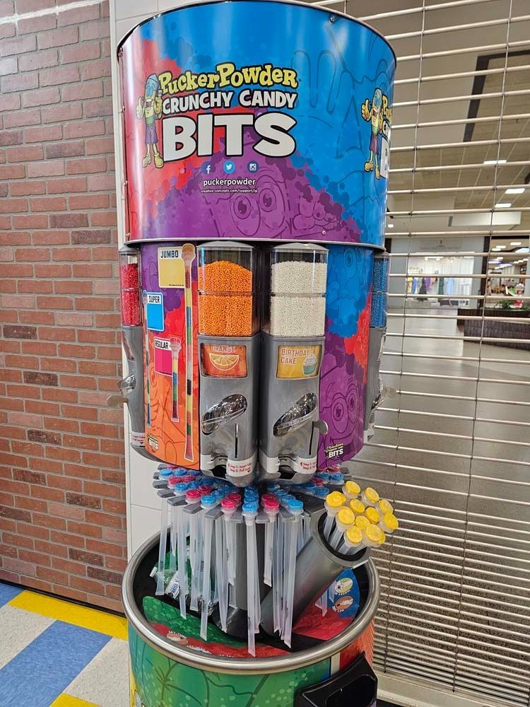 Colorful candy dispenser display in a brick-walled store, with packs of Bit O Honey Crunchy Candy Bits.