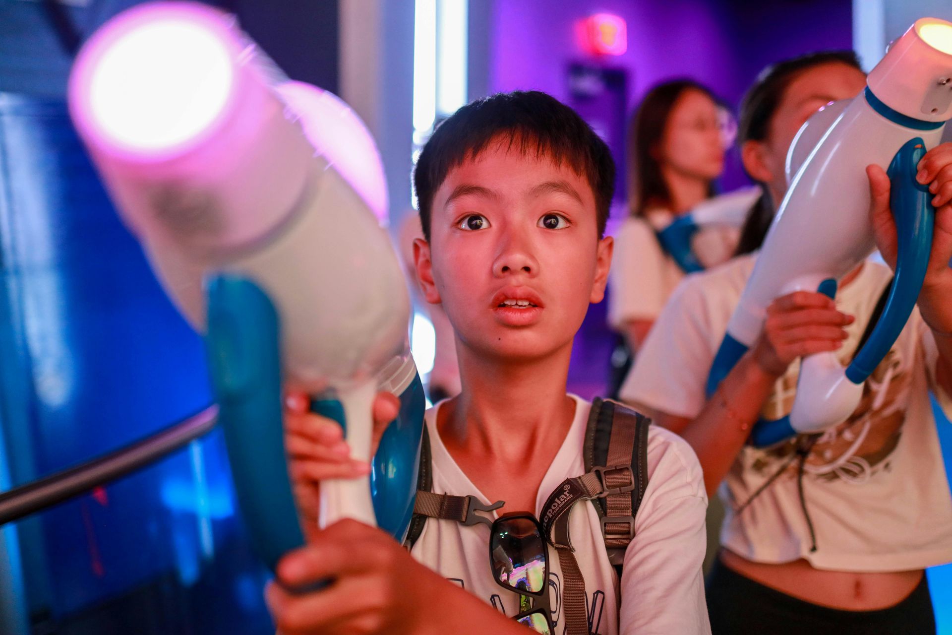 Boy holding a glowing toy wand at a colorful indoor event, looking surprised amid other attendees