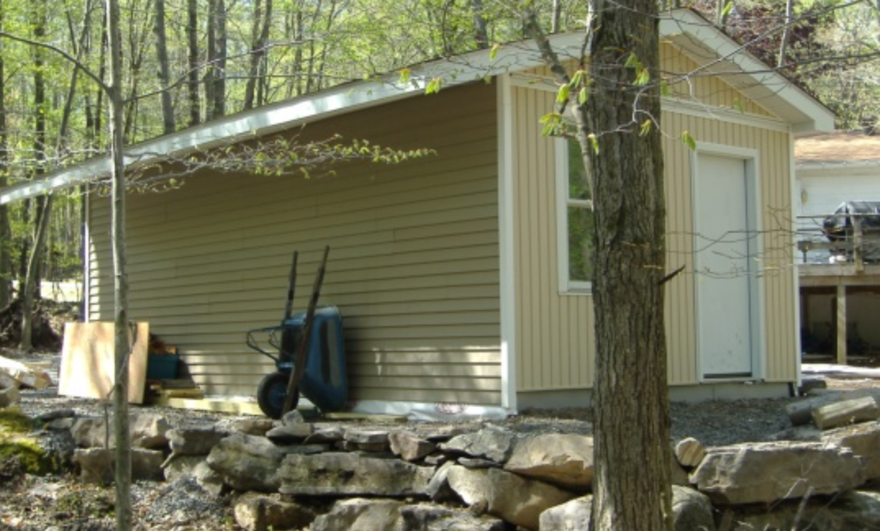 Small beige shed beside a stone wall in a wooded area, with a blue bin and a tree in front
