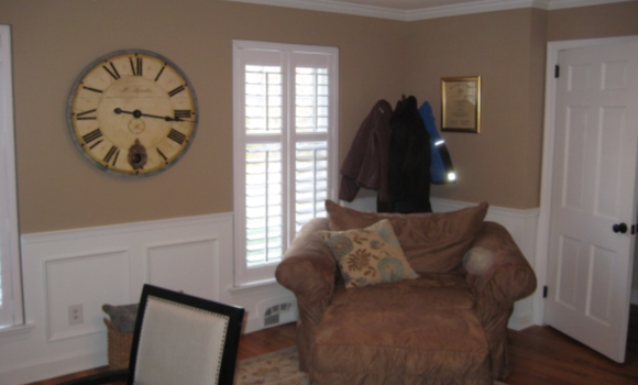 Cozy living room with armchair, wall clock, window shutters, and coat hooks on a tan wall