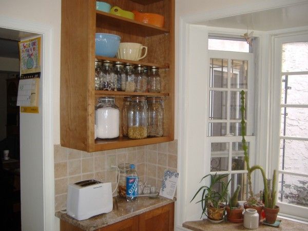 Kitchen corner with open wooden shelves, glassware, white toaster, and potted plants by a window
