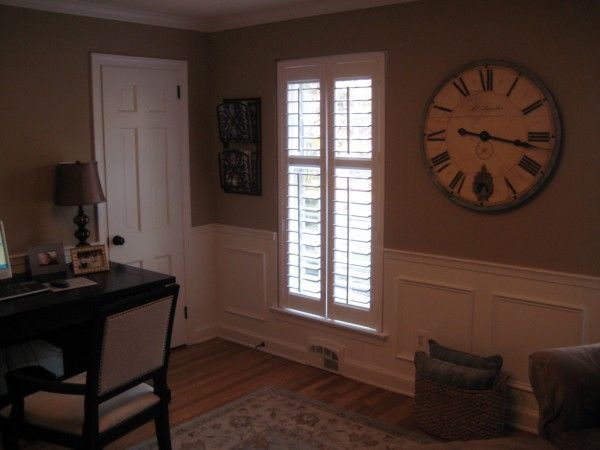Cozy home office with desk, lamp, window shutters, large wall clock, and basket on hardwood floor