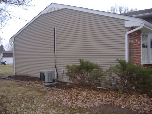 Side of beige house with an air conditioner unit, shrubs, and fallen leaves along the foundation.