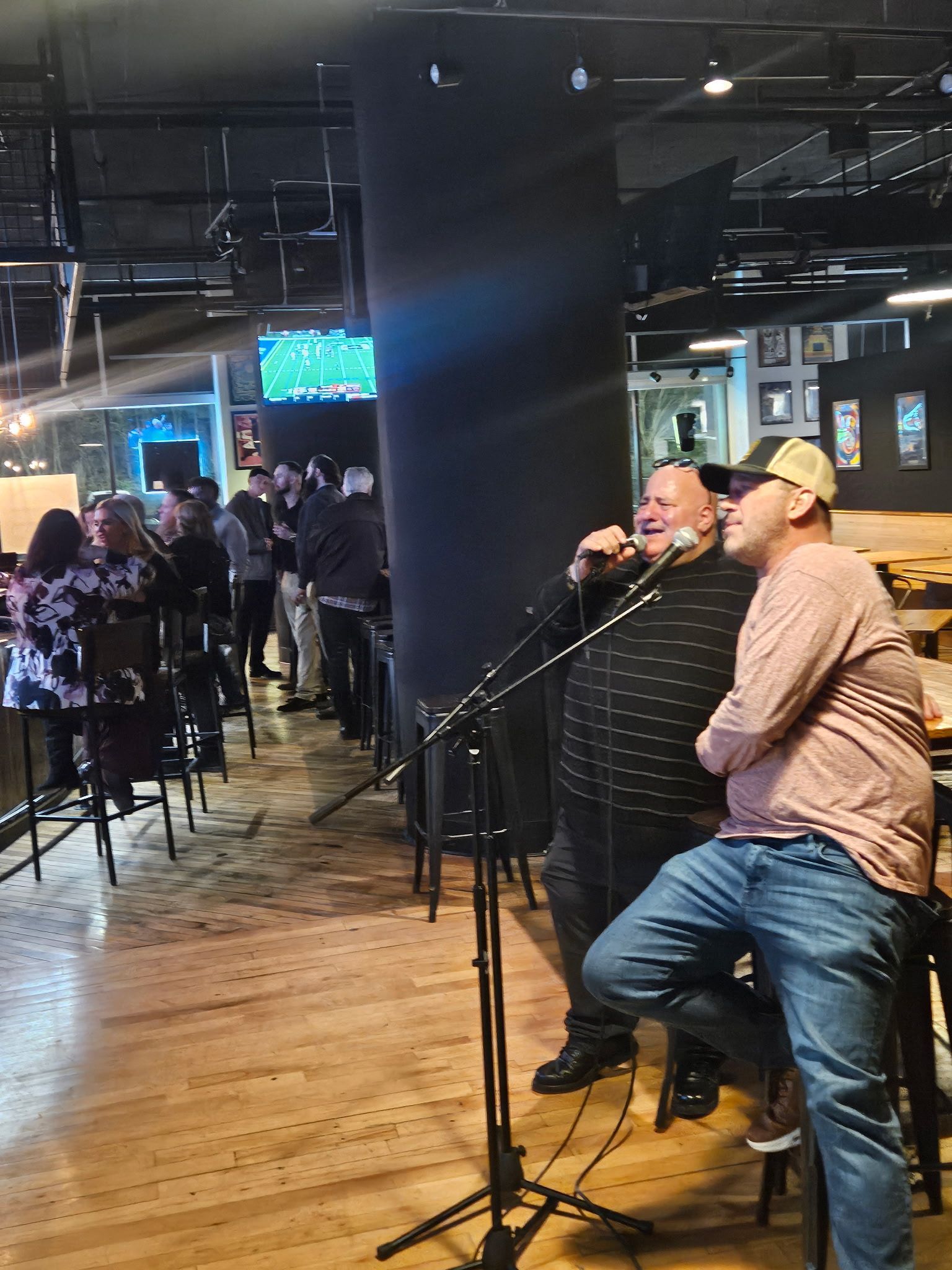 Two men singing karaoke at a bar with a microphone. People watch nearby.