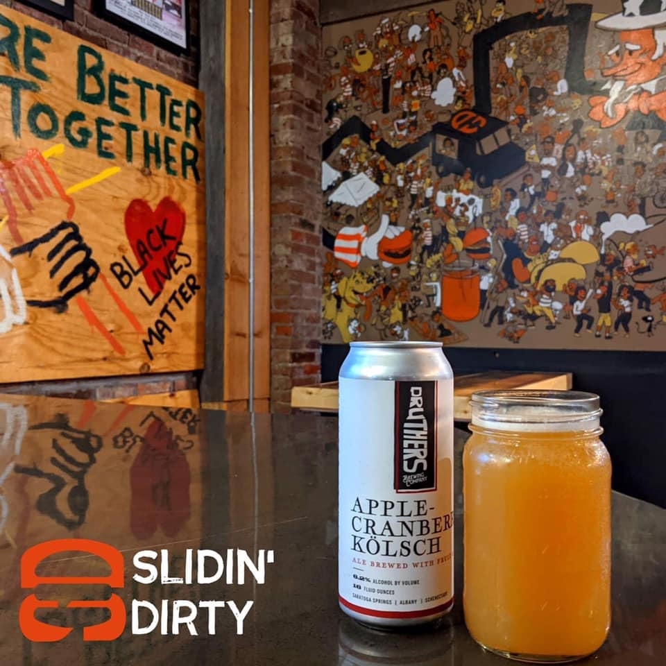 Can of apple-cranberry Kölsch and glass of orange liquid on a bar in front of graffiti art and a sign that says