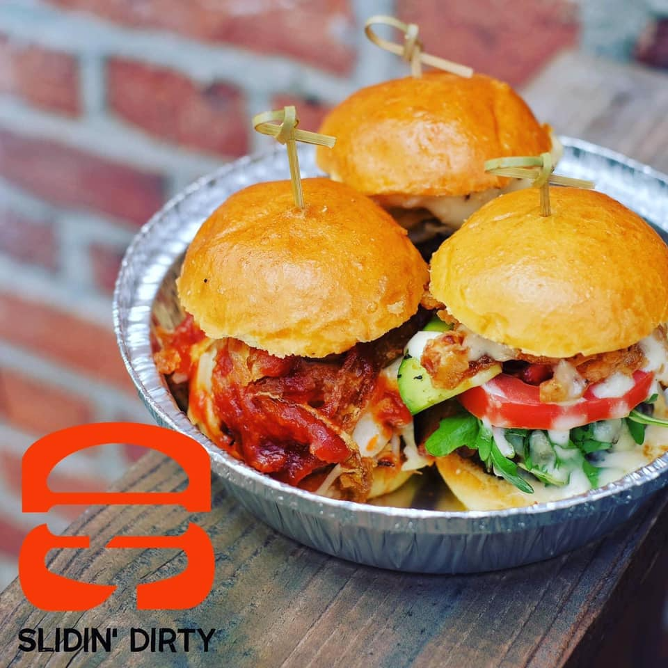 Three sliders in a foil dish; on a wooden surface in front of a brick wall. Logo for Slidin' Dirty.