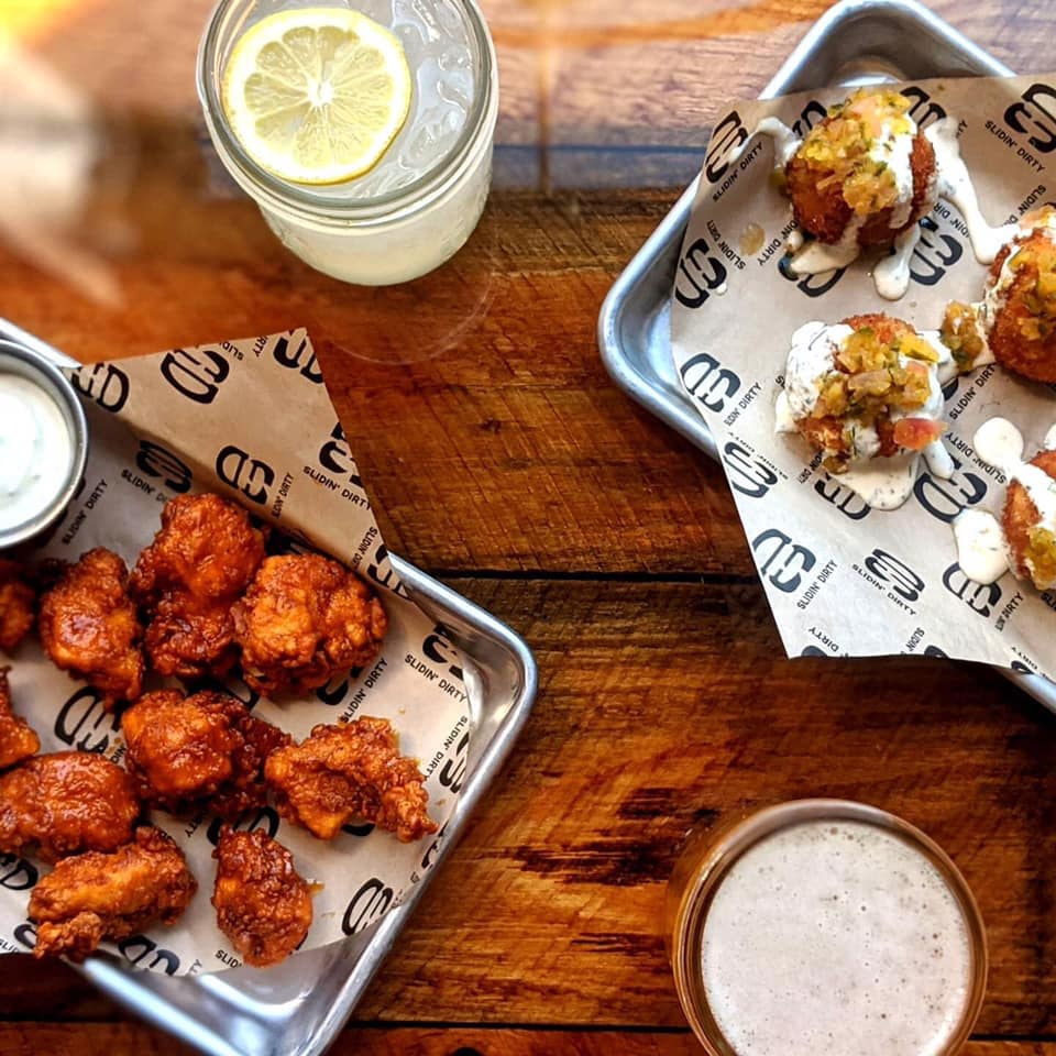 Platters of fried food with dipping sauces, drink with lemon slice, and beer on a wooden table.