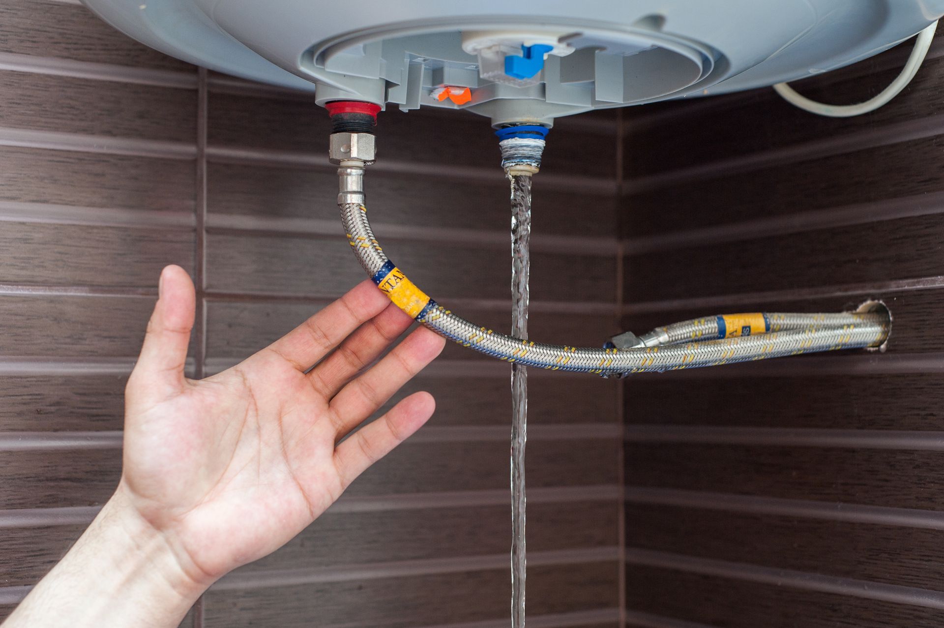 Water leaks from a water heater connection pipe while a hand reaches toward it against a dark tiled wall.