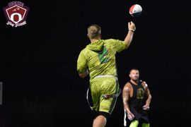 Flag football game at night, a player in red jersey runs with the ball as others pursue on a green field.