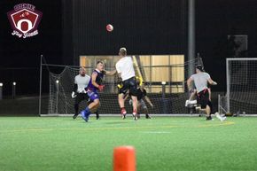 Flag football game at night, a player in red jersey runs with the ball as others pursue on a green field.