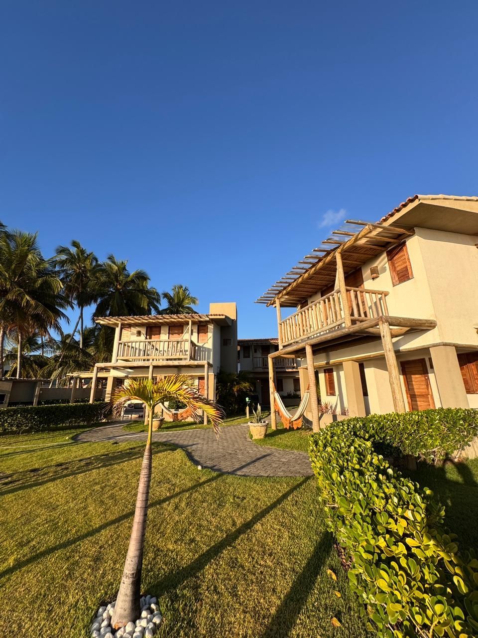 Duas casas de férias tropicais de dois andares com varandas de madeira, rodeadas por um gramado e palmeiras sob um céu azul brilhante.