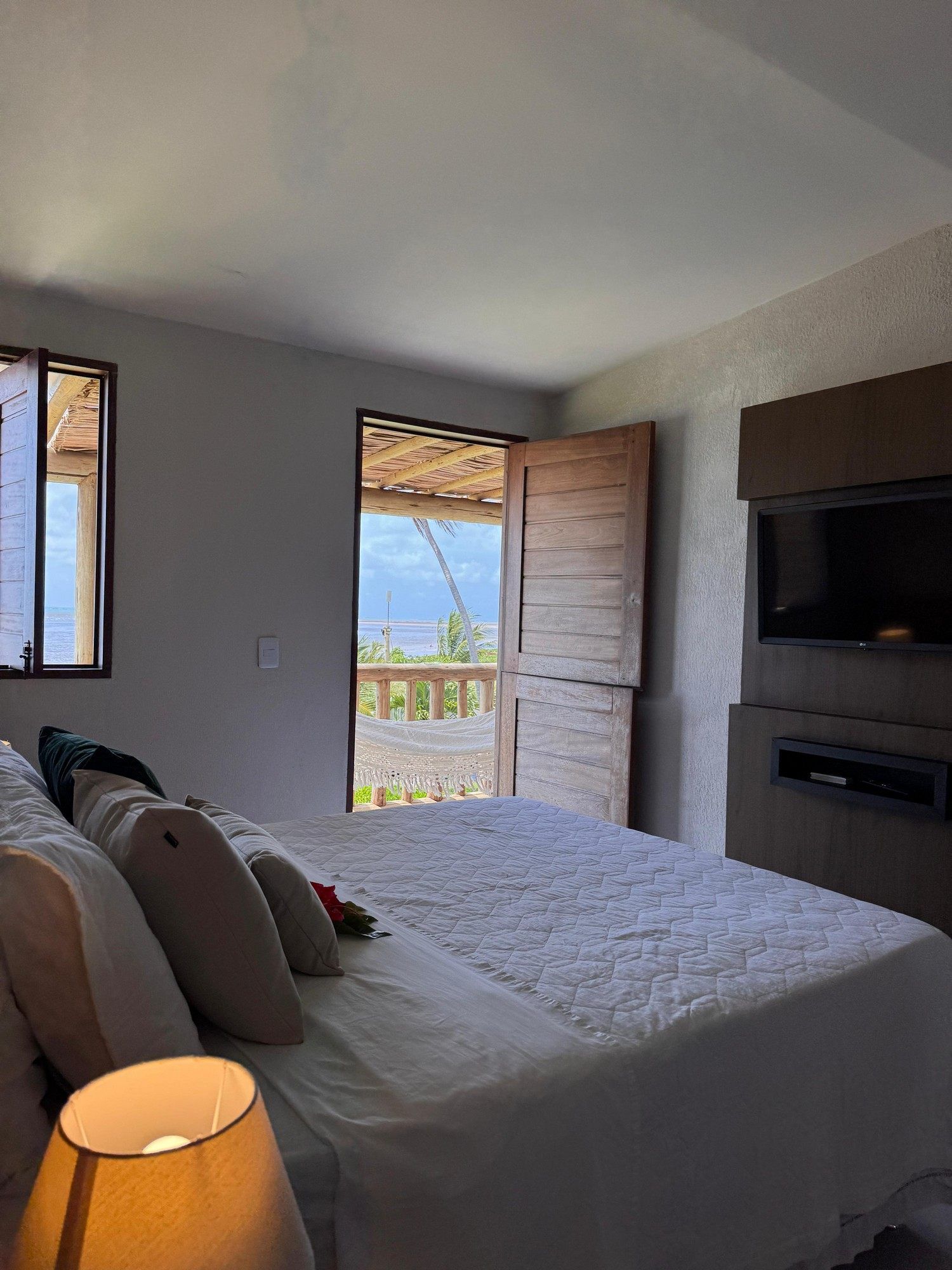 Um quarto aconchegante com uma cama branca macia, uma TV fixada na parede, um abajur e uma porta que se abre para um deck com vista panorâmica.