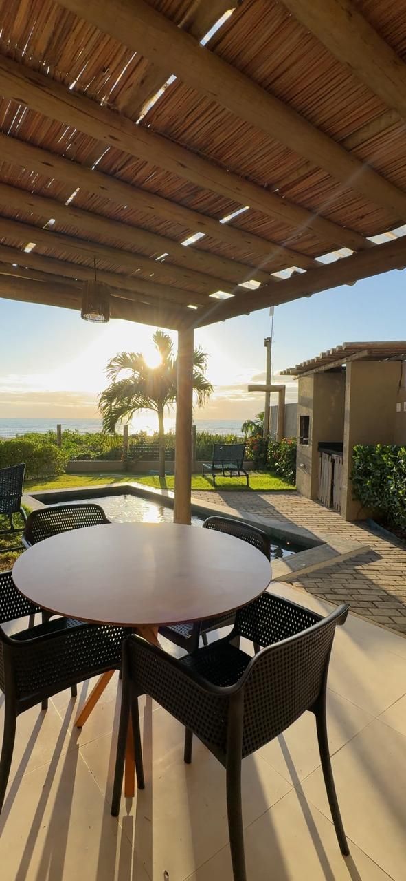 Uma mesa redonda com cadeiras fica sob uma pérgola de madeira em um pátio, com vista para uma pequena piscina, uma palmeira e o pôr do sol no oceano.