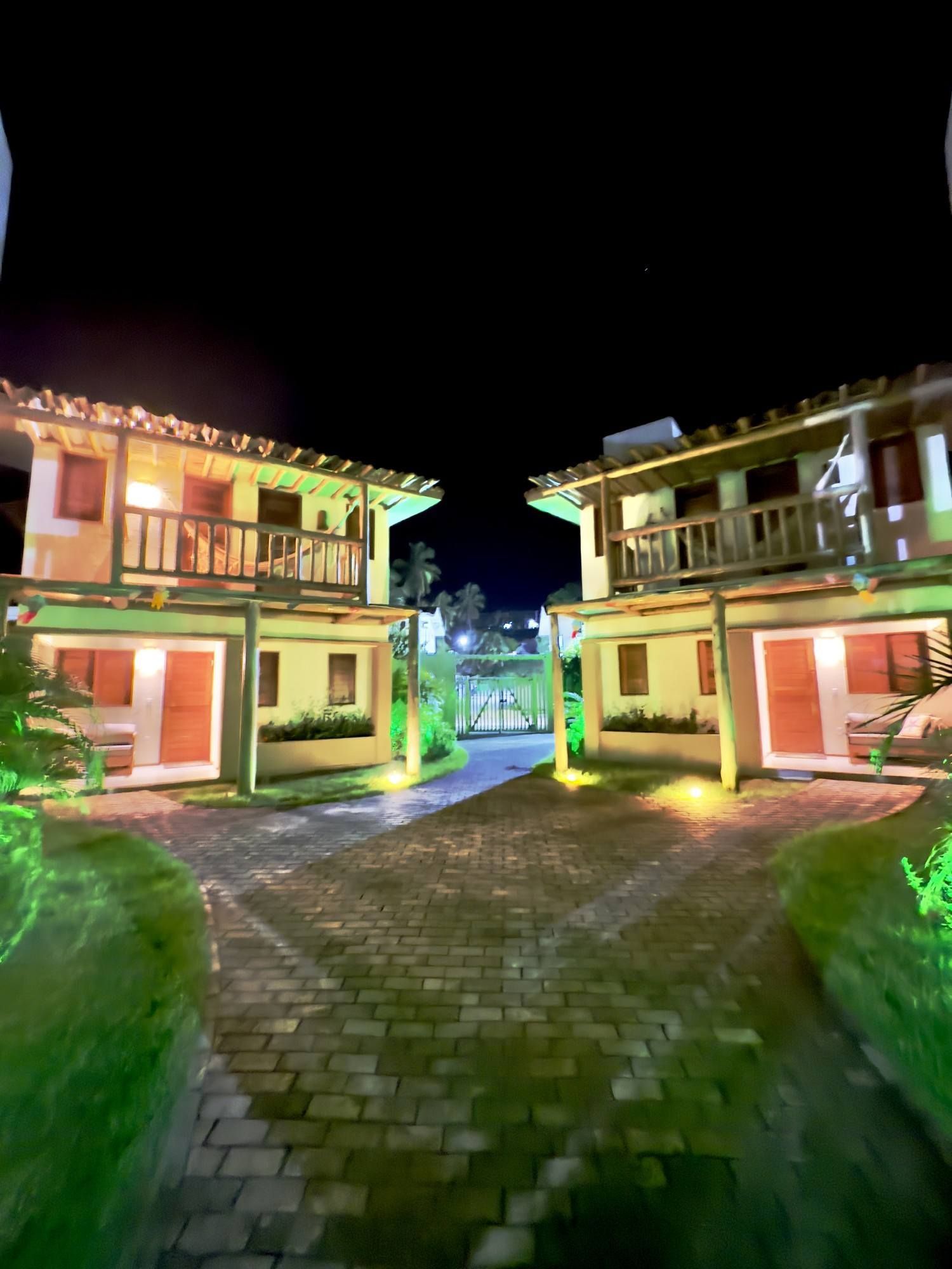 Dois edifícios de dois andares com varandas de madeira, iluminados por luzes aconchegantes à noite, ladeiam um caminho de paralelepípedos em um pátio ajardinado.