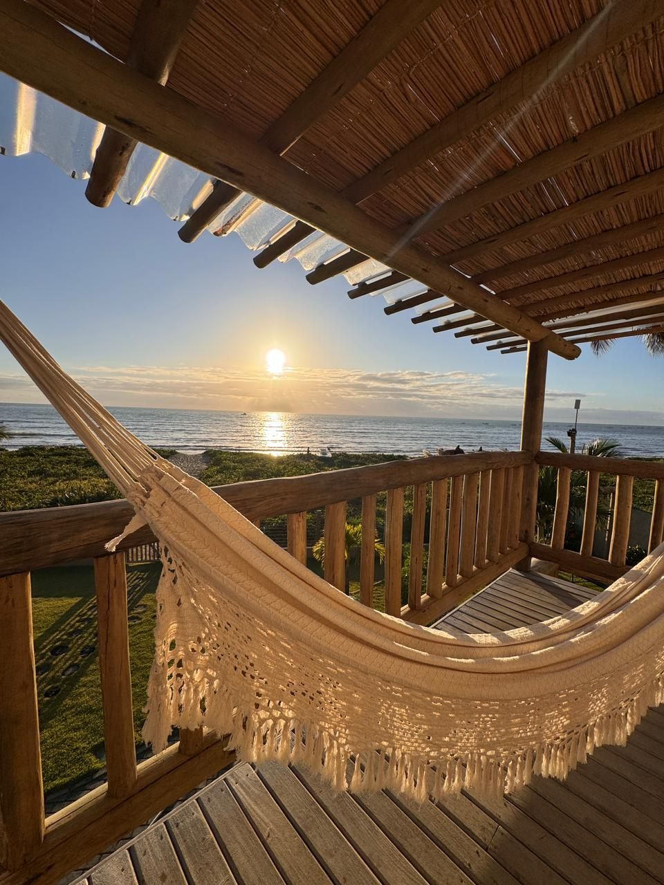 Uma rede cor creme em um deck de madeira com vista para a praia ao pôr do sol, sob uma estrutura com teto de palha.